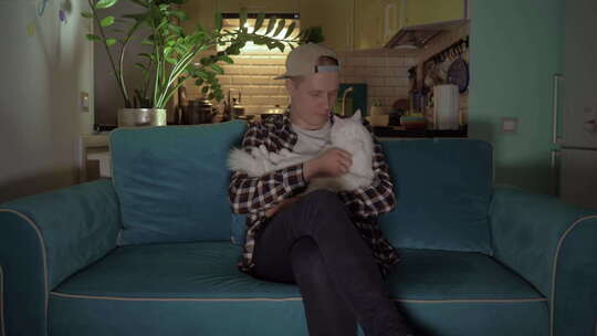 这家伙坐在蓝色沙发上，抚摸着一只躺在膝盖上的白猫