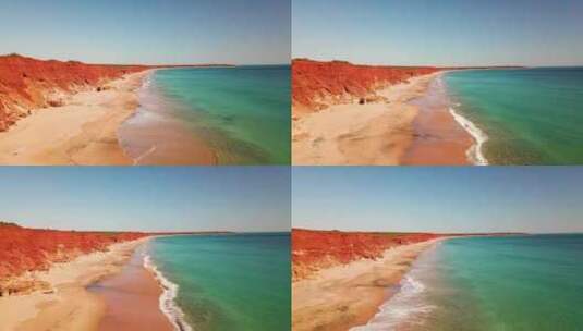 无人机在沙滩上的红色悬崖旁飞翔，碧蓝色的水，海浪在下面涌动高清在线视频素材下载