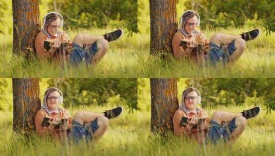 戴着眼镜，戴着耳机看书的年轻人，夏天坐在树旁。高清在线视频素材下载