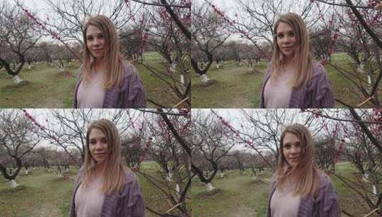 年轻敏感的女人带着可爱的微笑和自然的妆容，在刮风的天气里站在樱花盛开的树枝上。4k镜头。高清在线视频素材下载