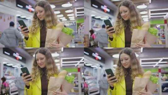 年轻女子在杂货店使用智能手机，纸袋里装着高清在线视频素材下载