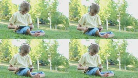 男孩戴着耳机坐在户外草地上玩平板电脑高清在线视频素材下载